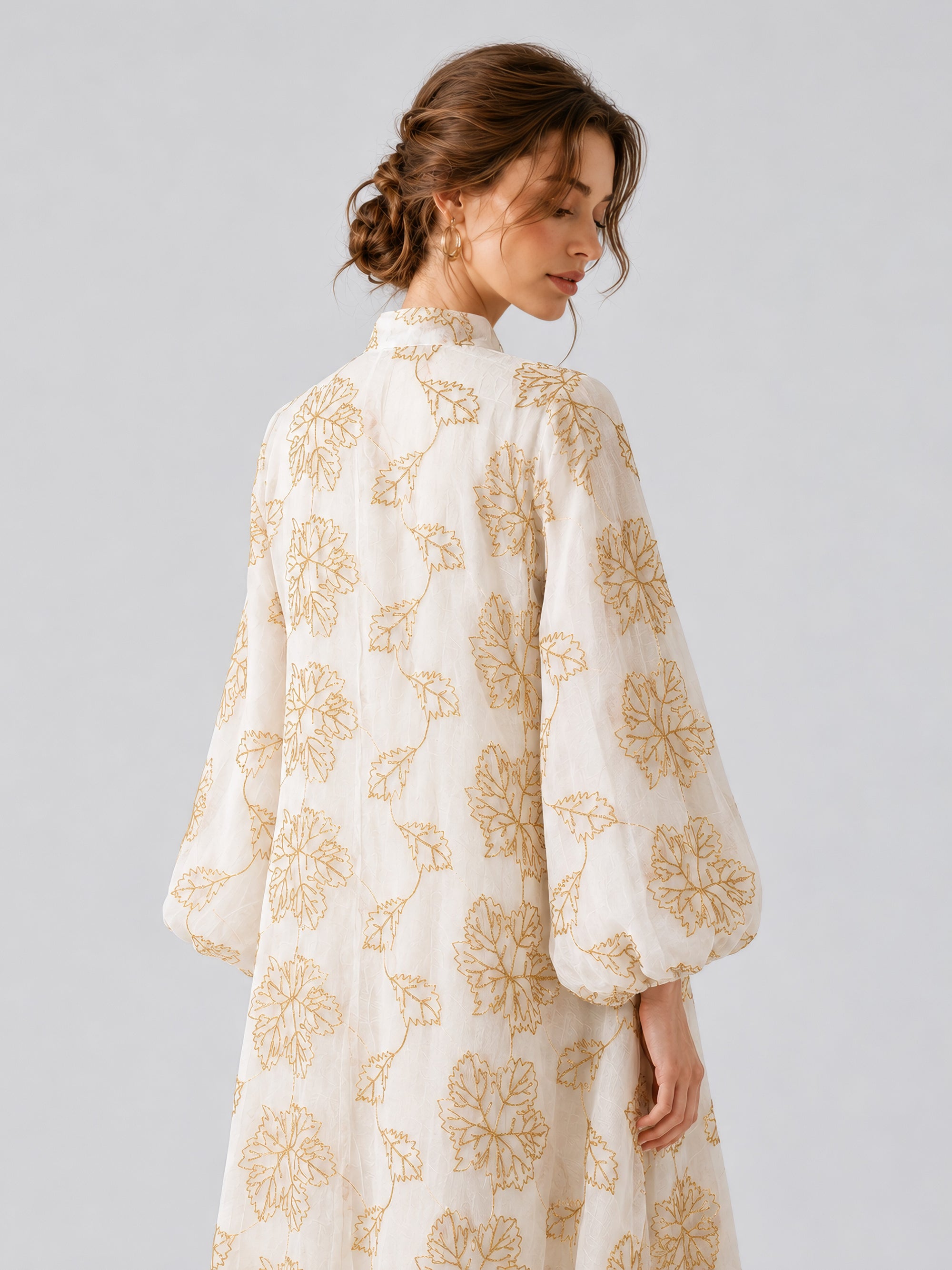 A woman wearing the Social Queen Leaf Embroidered Beaded Dress, featuring gold floral patterns, stands with her back to the camera and her head turned to the side against a plain light background.