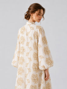 A woman wearing the Social Queen Leaf Embroidered Beaded Dress, featuring gold floral patterns, stands with her back to the camera and her head turned to the side against a plain light background.