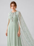 A woman models the Social Queen Beaded Printed Cape Chiffon Dress, featuring jeweled embellishments and a lightweight green chiffon maxi silhouette with a sheer, printed cape overlay, set against a plain background.