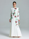 A woman stands against a plain light background wearing the Social Queen Floral Beaded A-Line Gown, featuring long sleeves and pink and green accents, and holds a small handbag.