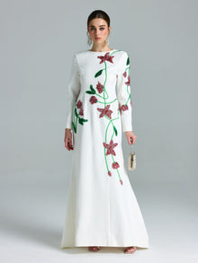 A woman stands against a plain light background wearing the Social Queen Floral Beaded A-Line Gown, featuring long sleeves and pink and green accents, and holds a small handbag.