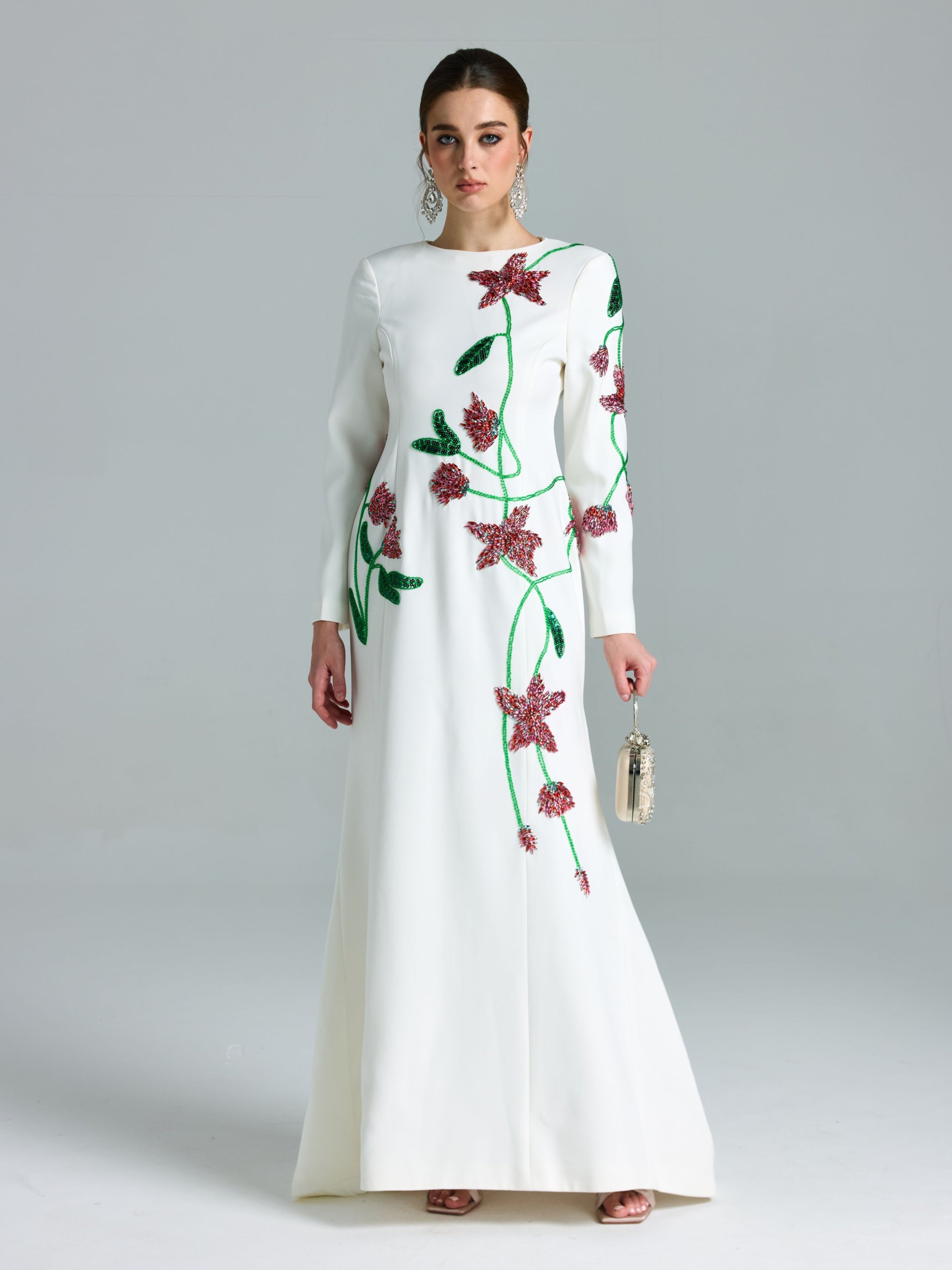 A woman stands against a plain light background wearing the Social Queen Floral Beaded A-Line Gown, featuring long sleeves and pink and green accents, and holds a small handbag.