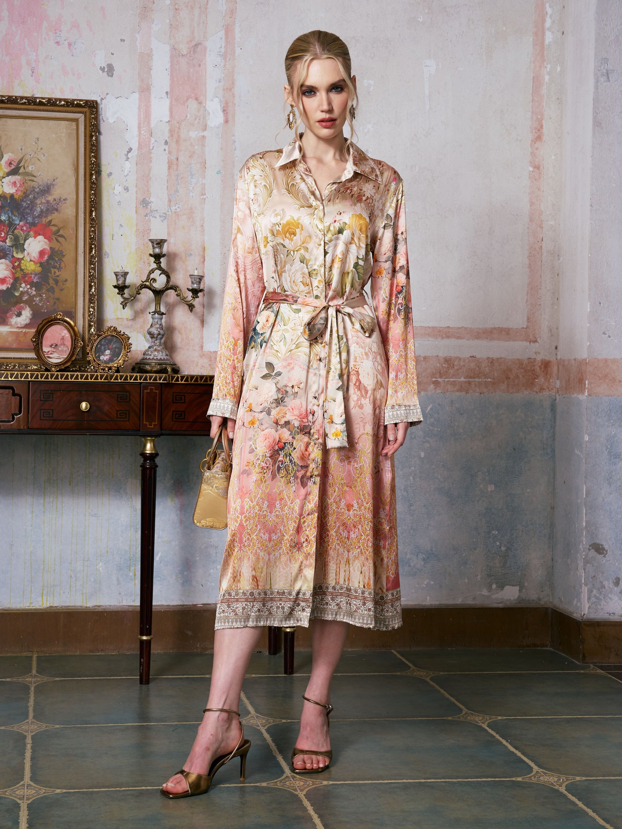 A woman stands indoors on a tiled floor, holding a small handbag, wearing the Garden Print Mulberry Silk Shirt Dress by Social Queen—a long, belted midi dress in a baroque garden print—with artwork and decor arranged on a table behind her.