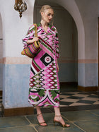 A woman wearing the Social Queen Geometric Print Mulberry Silk Dress and holding a colorful woven bag stands indoors on tiled flooring beneath an arched doorway.
