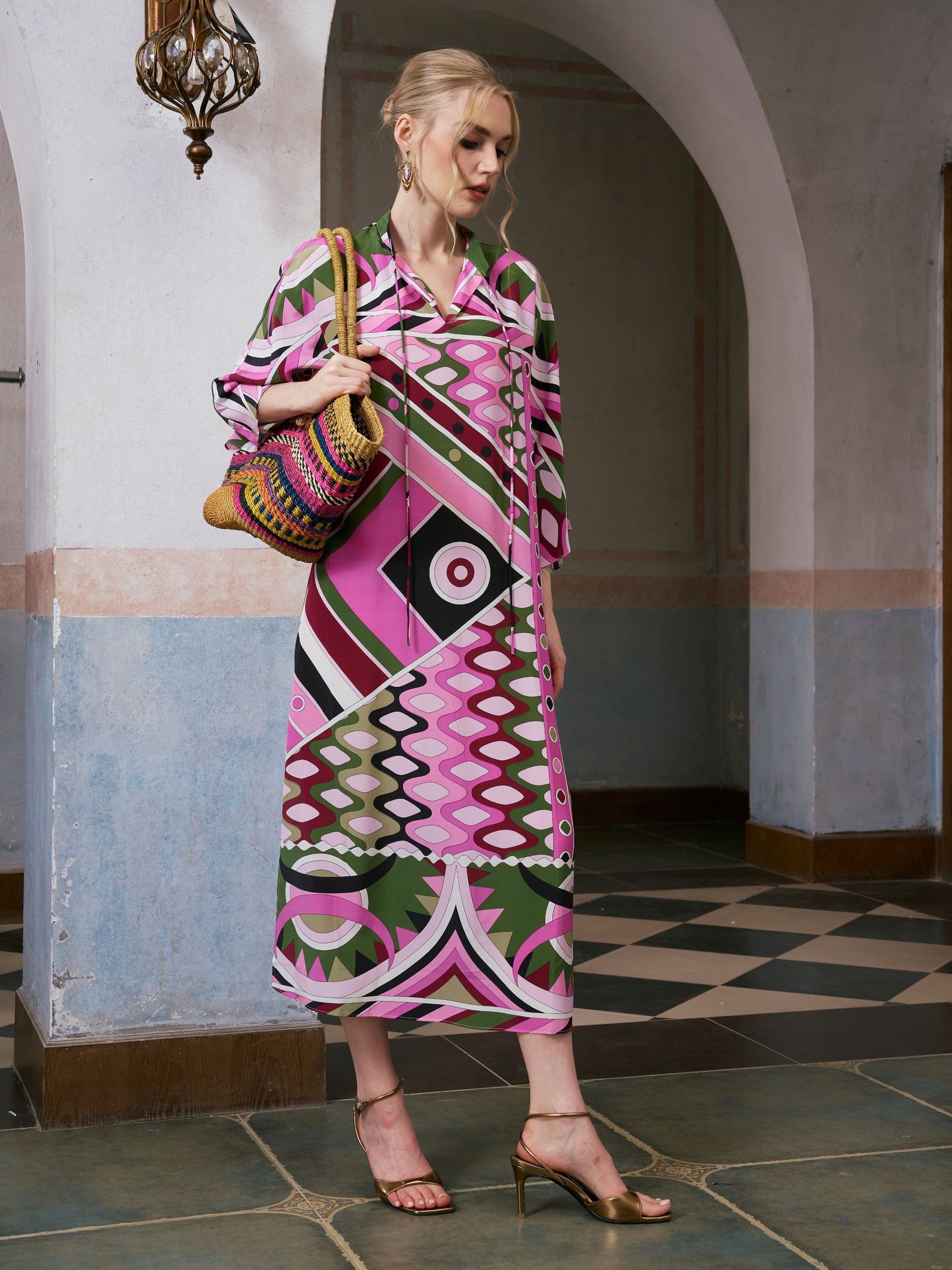 A woman wearing the Social Queen Geometric Print Mulberry Silk Dress and holding a colorful woven bag stands indoors on tiled flooring beneath an arched doorway.