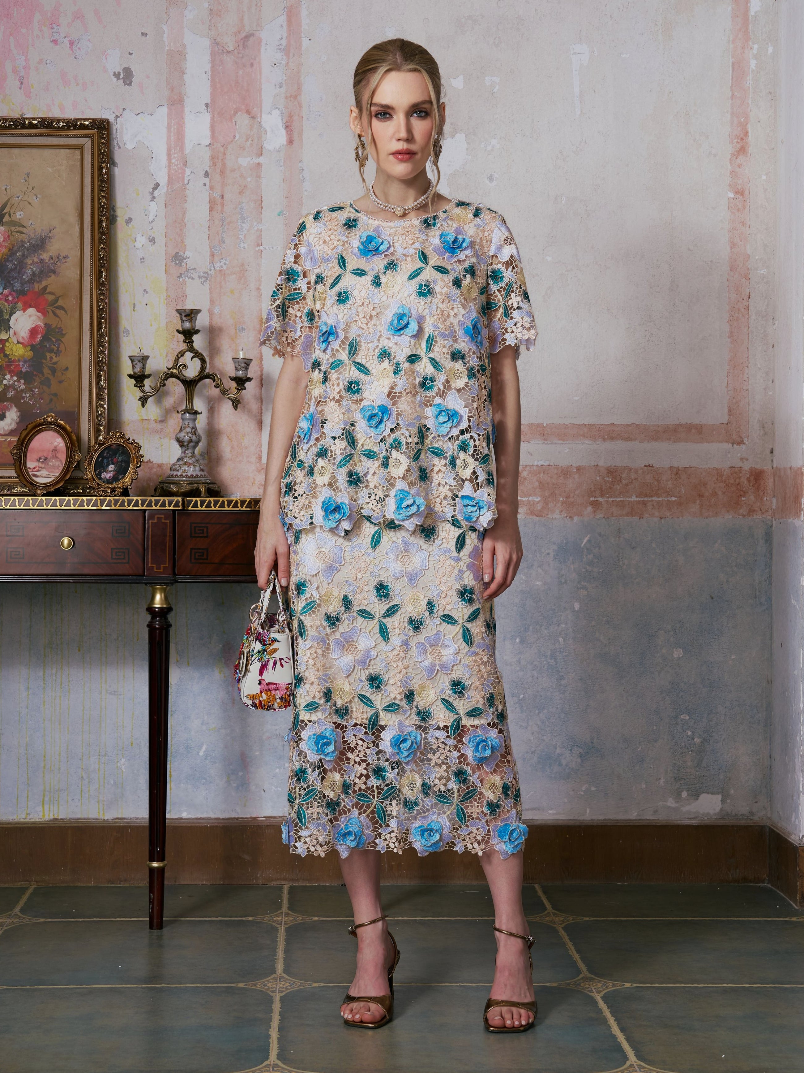 A woman stands indoors wearing the Social Queen 3D Floral Cutwork Lace Top, styled with a matching skirt in blue and beige patterns featuring delicate floral embroidery, holding a small handbag, with a decorated table and framed painting in the background.