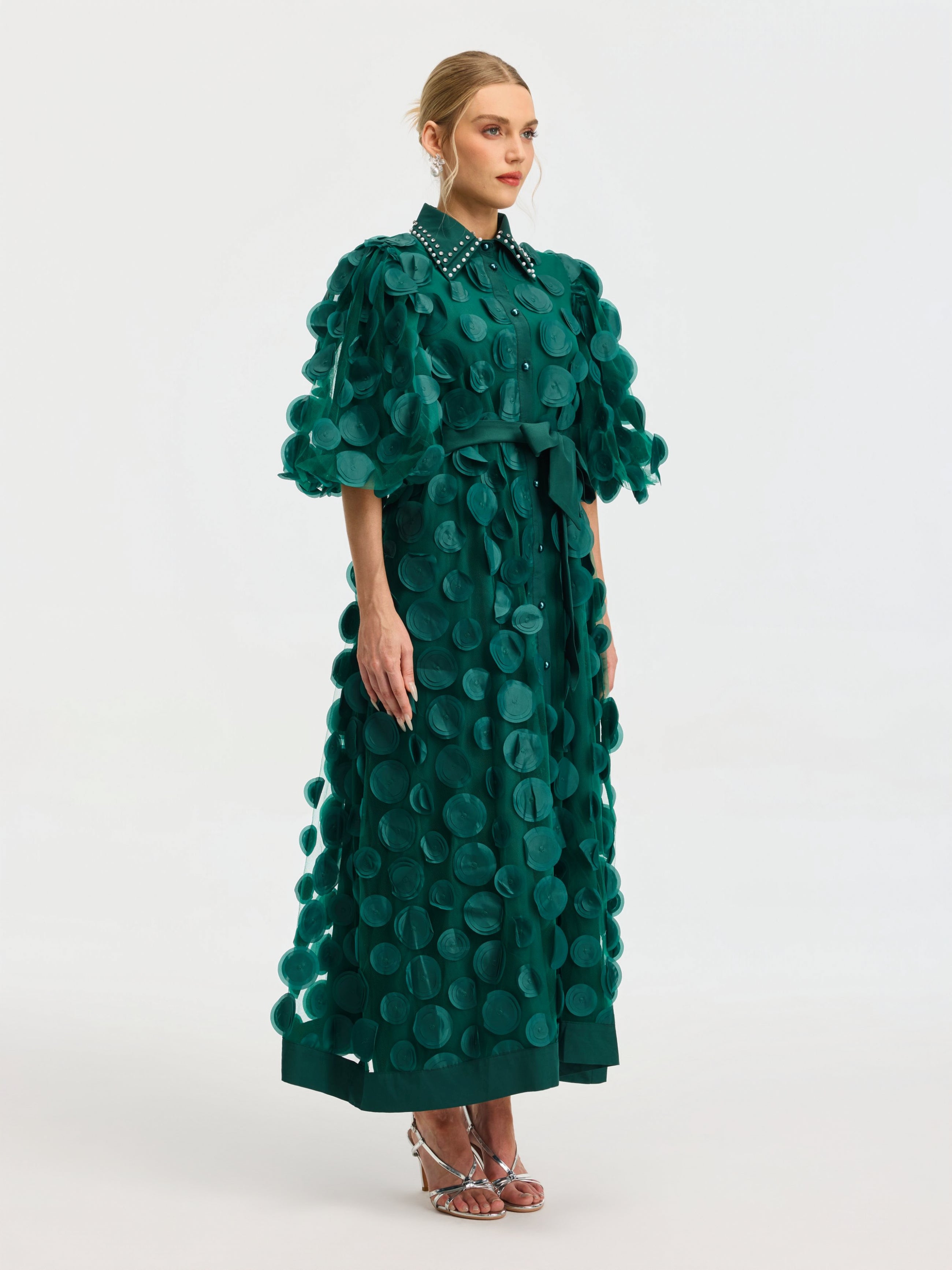 A woman wears the Social Queen Pearl Embellished Appliqué Dress—an ankle-length green A-line with short sleeves, belted waist, and large fabric appliqués—paired with silver strappy sandals against a plain background.