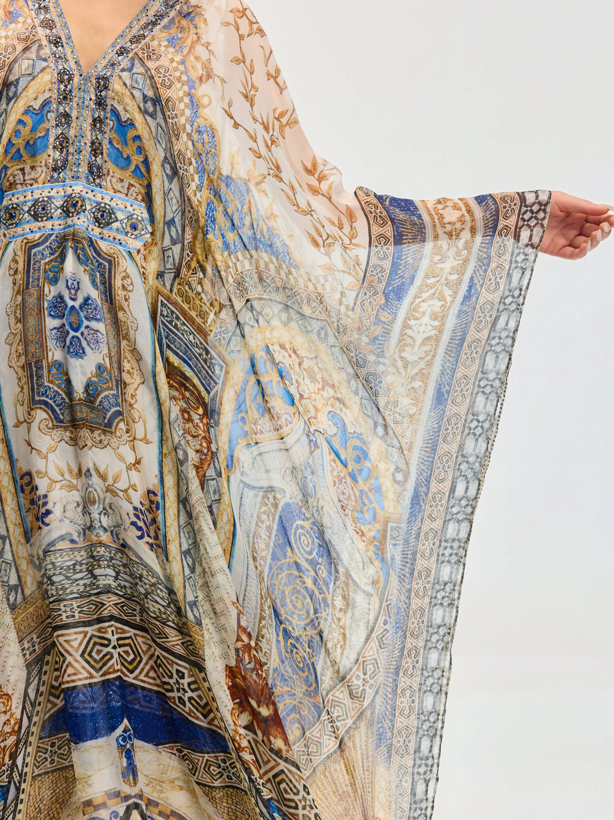 A person showcases the Social Queen Byzantine Print Silk Caftan Dress, featuring blue, gold, and beige intricate designs and a crystal-embellished neckline, extending one arm to reveal its wide sleeve against a plain white background.