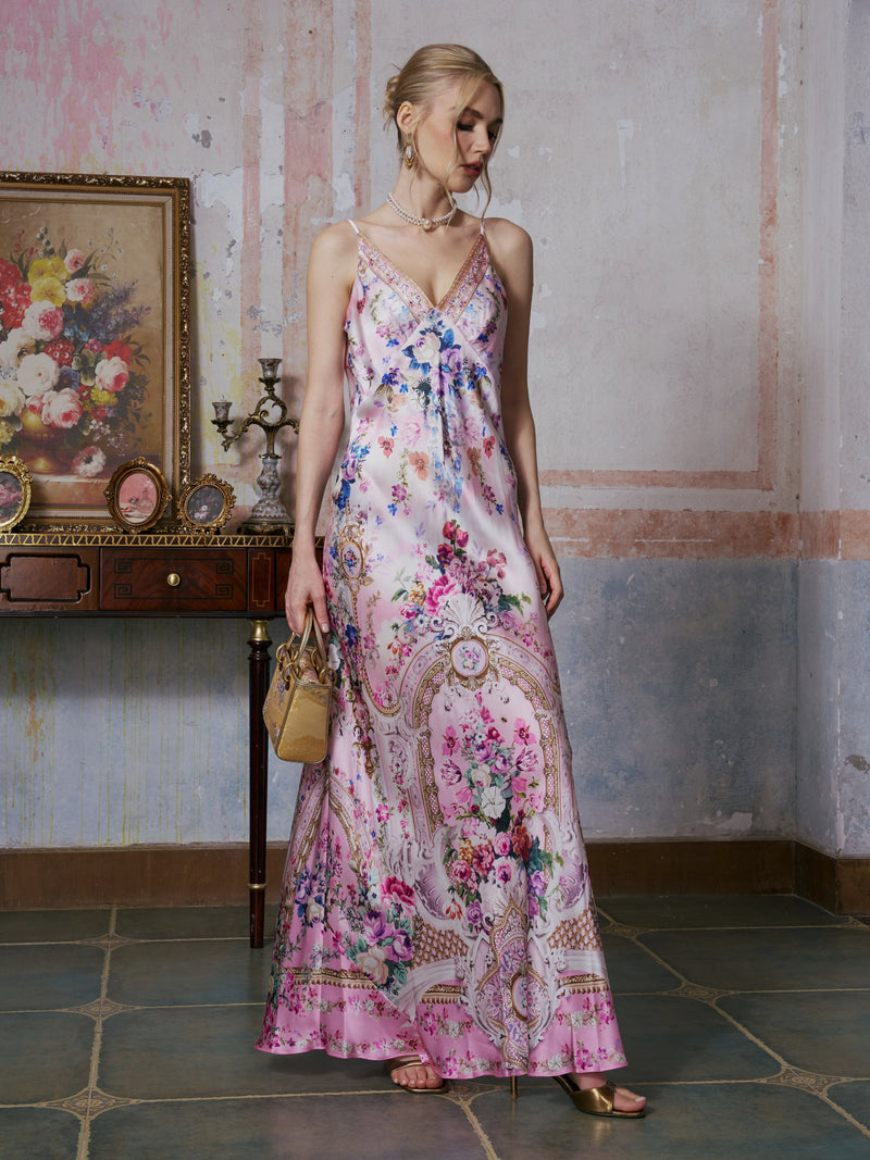 A woman stands indoors holding a small beige handbag, wearing the Baroque Print Mulberry Silk Dress by Social Queen. The background includes a vintage table, framed floral painting, and candelabra.