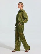 A woman in profile wears Social Queen's Botanical Burnout Satin Wide-Leg Pants—a loose, sheer green style with a subtle floral pattern—paired with open-toe sandals, standing against a plain light background.