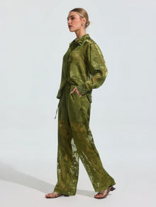 A woman in profile wears Social Queen's Botanical Burnout Satin Wide-Leg Pants—a loose, sheer green style with a subtle floral pattern—paired with open-toe sandals, standing against a plain light background.