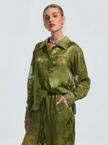 A woman wears the Social Queen Beaded Botanical Burnout Satin Shirt in olive green with matching pants, featuring sheer botanical texture and beaded lace trim, paired with a pearl necklace against a plain light background.
