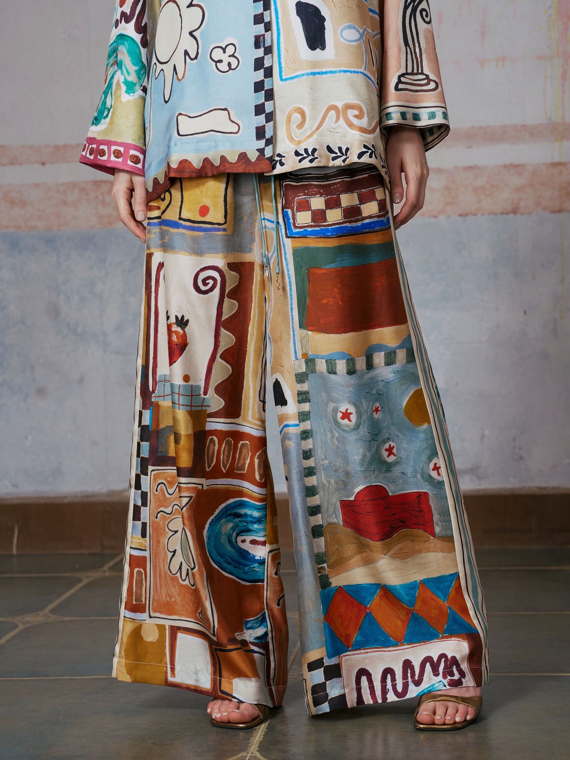A person wearing the Social Queen Abstract Print Mulberry Silk Pants and a matching top, both showcasing colorful, abstract geometric and nature-inspired patterns, stands indoors on a tiled floor.