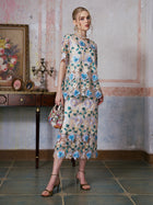 A woman stands indoors wearing a floral lace top and the Social Queen 3D Floral Cutwork Lace Skirt with blue flowers, holding a patterned handbag beside an antique table adorned with a framed floral painting and decorative items.