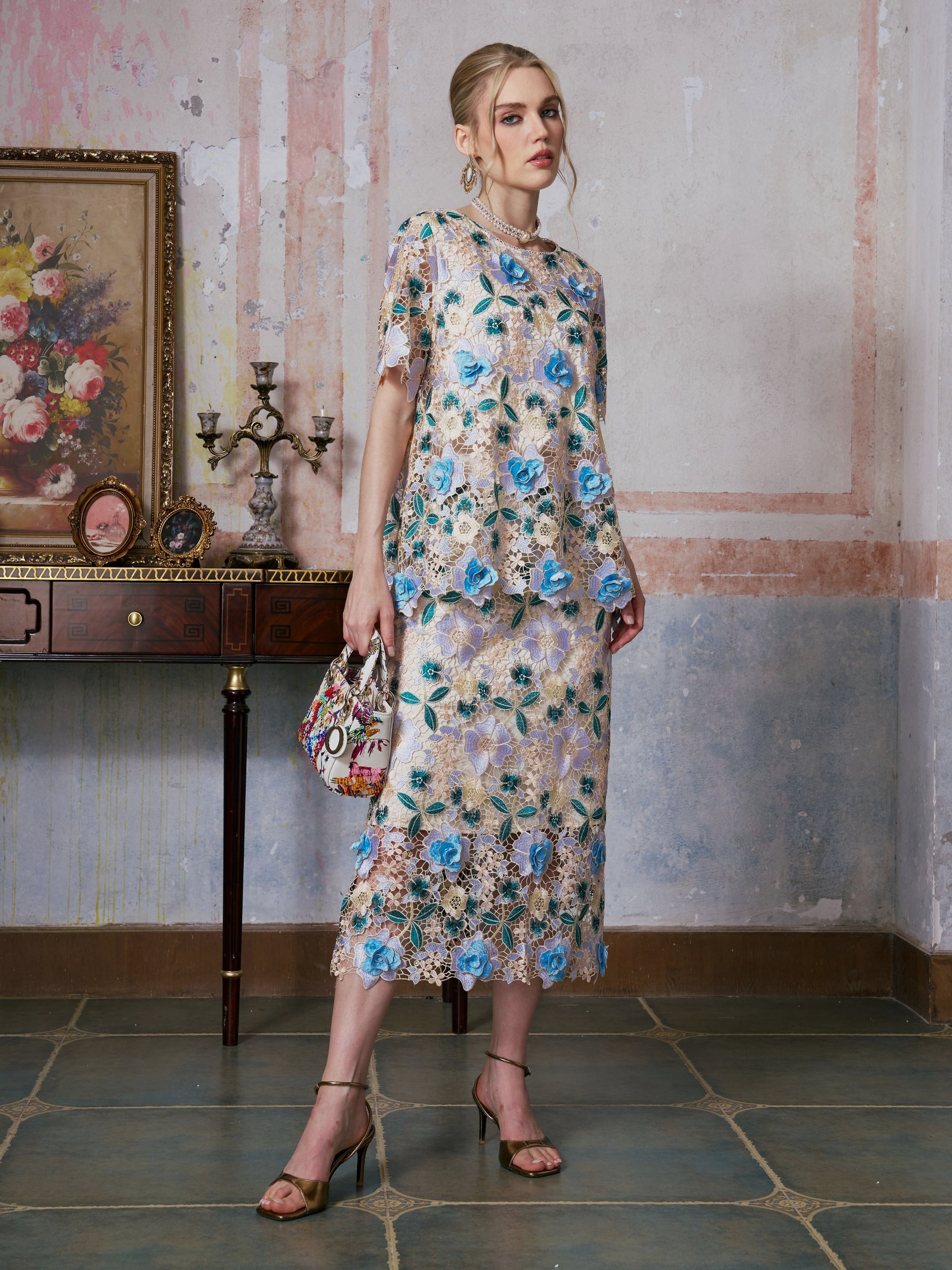 A woman stands indoors wearing a floral lace top and the Social Queen 3D Floral Cutwork Lace Skirt with blue flowers, holding a patterned handbag beside an antique table adorned with a framed floral painting and decorative items.