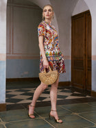 A woman in Social Queen’s Paisley Garden Print Silk Shorts and gold heels holds a bamboo handbag while walking indoors on a tiled floor near wooden doors and arched walls.