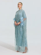 Wearing the Social Queen Beaded Embroidered Feather Trim Cape Gown—a light blue, floor-length dress with sheer overlay and feathered sleeves—this elegant gown makes a statement against a simple background.