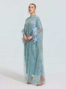 Wearing the Social Queen Beaded Embroidered Feather Trim Cape Gown—a light blue, floor-length dress with sheer overlay and feathered sleeves—this elegant gown makes a statement against a simple background.