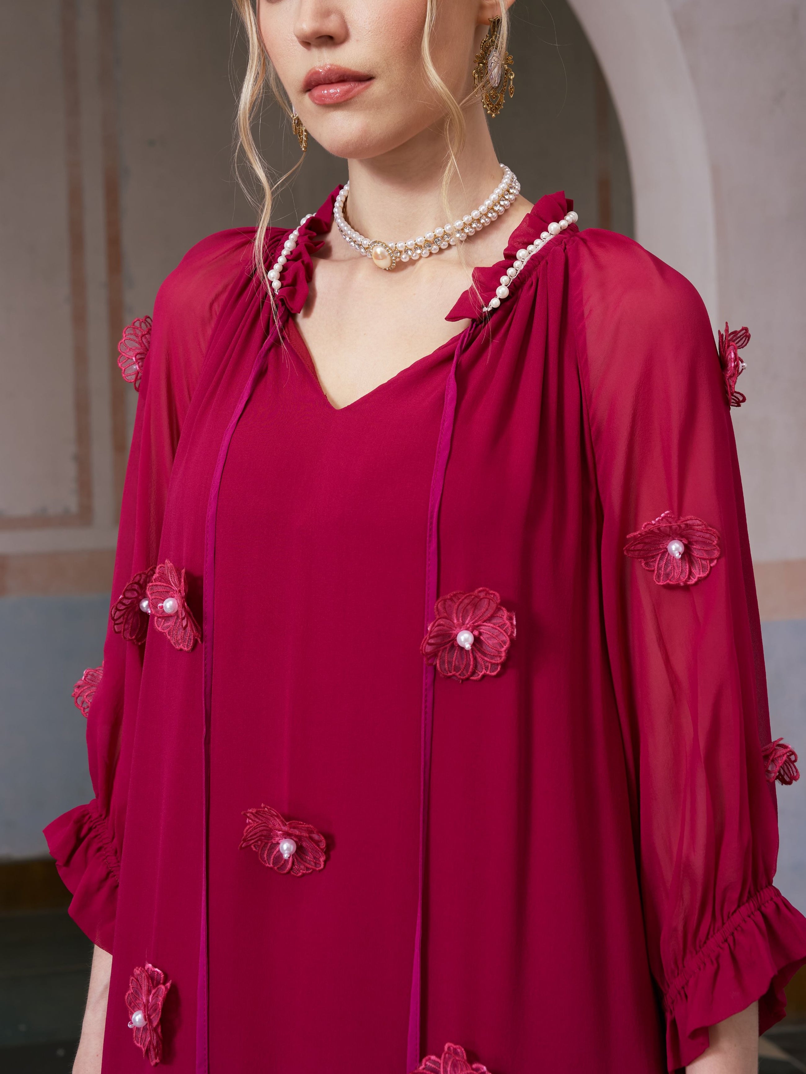 A woman wears the Social Queen 3D Floral Pearl Mulberry Silk Dress, featuring sheer, deep pink fabric with intricate floral appliqués. She accessorizes with an exquisite pearl necklace and gold earrings, while only her upper body and part of her face are visible.