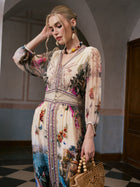 A woman stands indoors against muted walls and a wooden door, exuding effortless resort-wear elegance in the Social Queen Coastal Print Mulberry Silk Dress. She holds a bamboo handbag and touches her head, showcasing the dress’s striking Mediterranean-inspired pattern.
