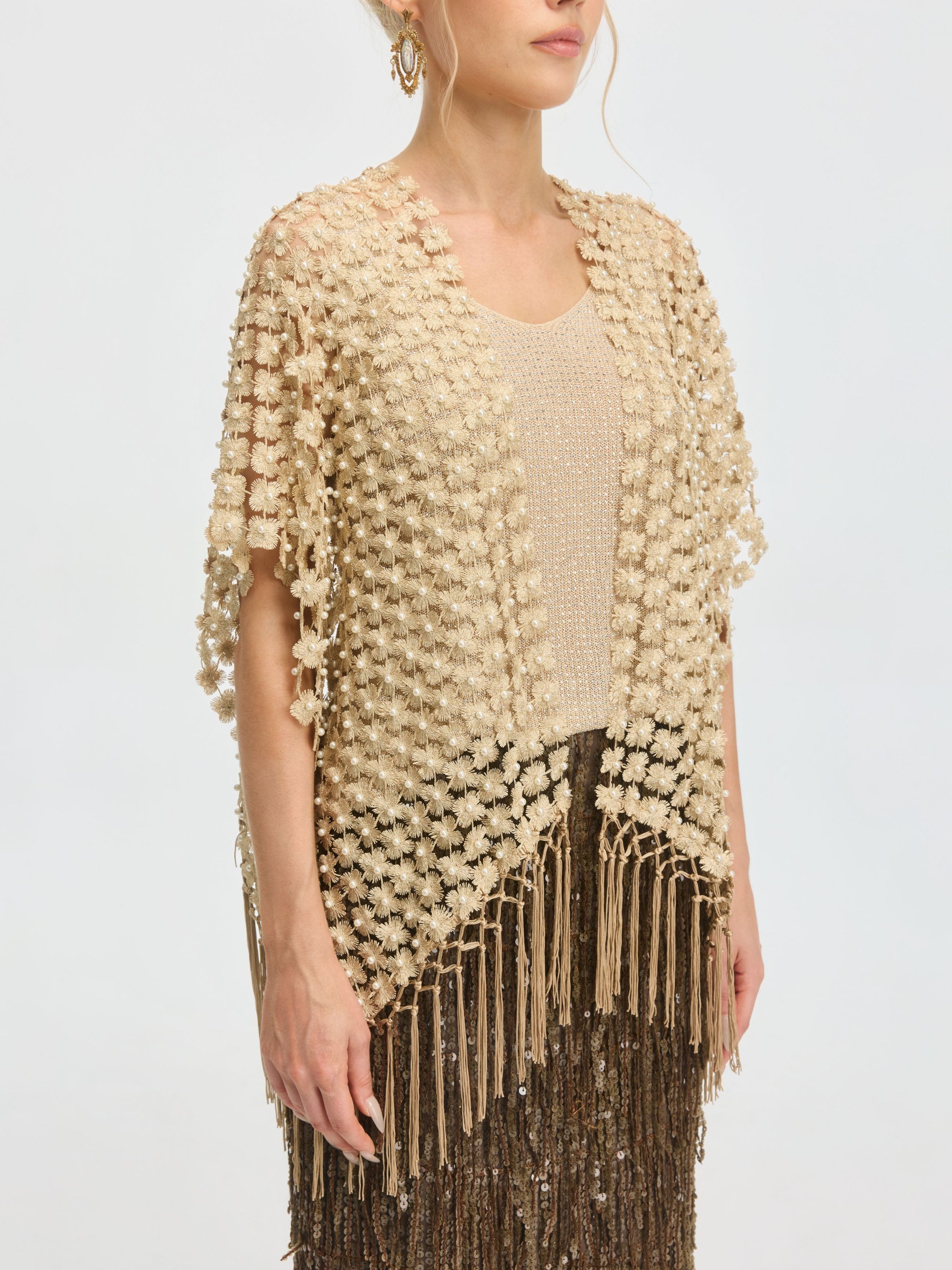 A woman wears the Social Queen Pearl Floral Appliqué Fringe Cape—a beige, textured, short-sleeve evening piece with fringe and floral details—over a dark sequined skirt.