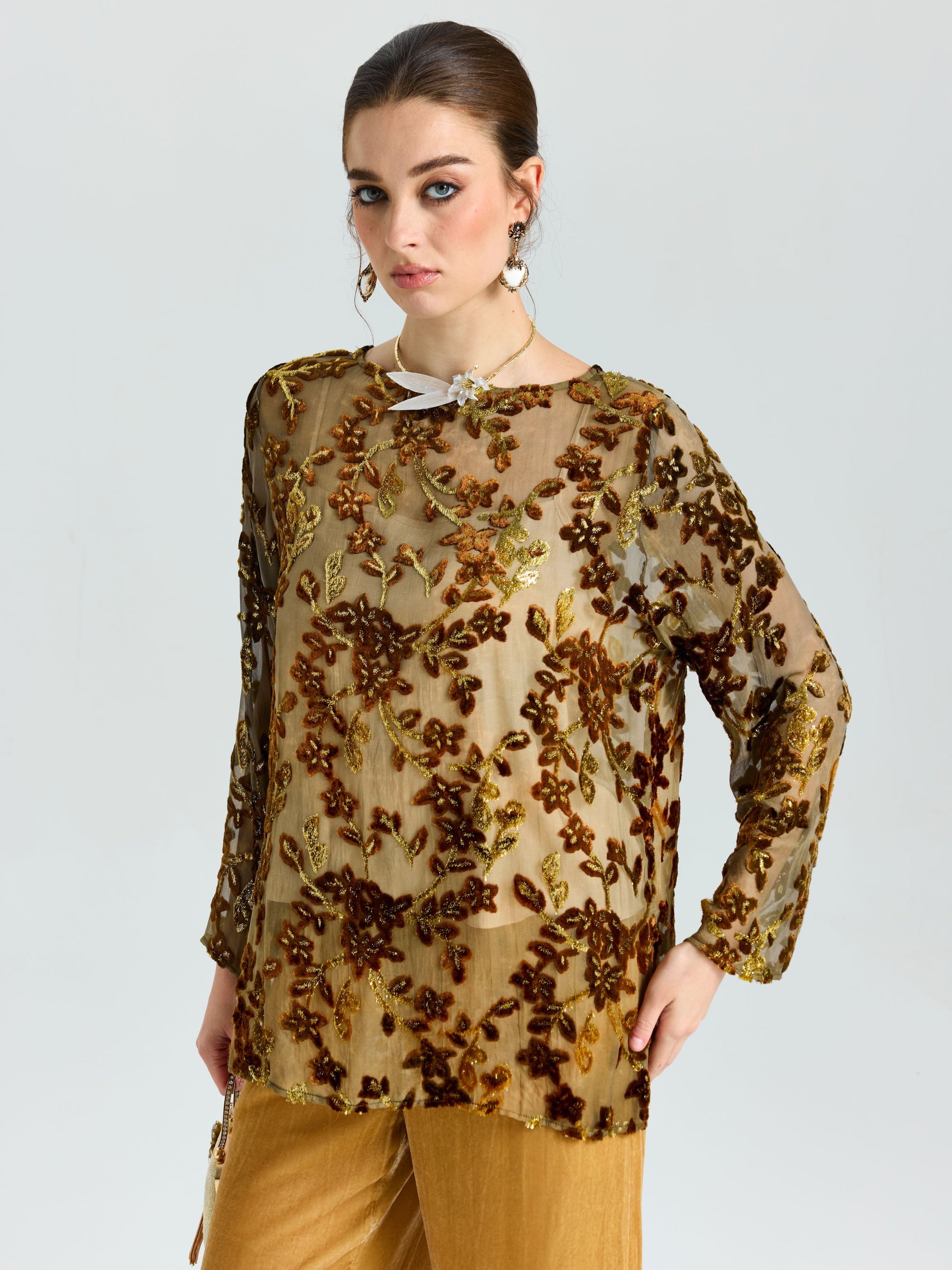 A woman wears the Social Queen Flocked Floral Silk Sheer Top (camisole not included), featuring metallic vine-inspired embroidery, paired with gold pants, standing against a plain light gray background.