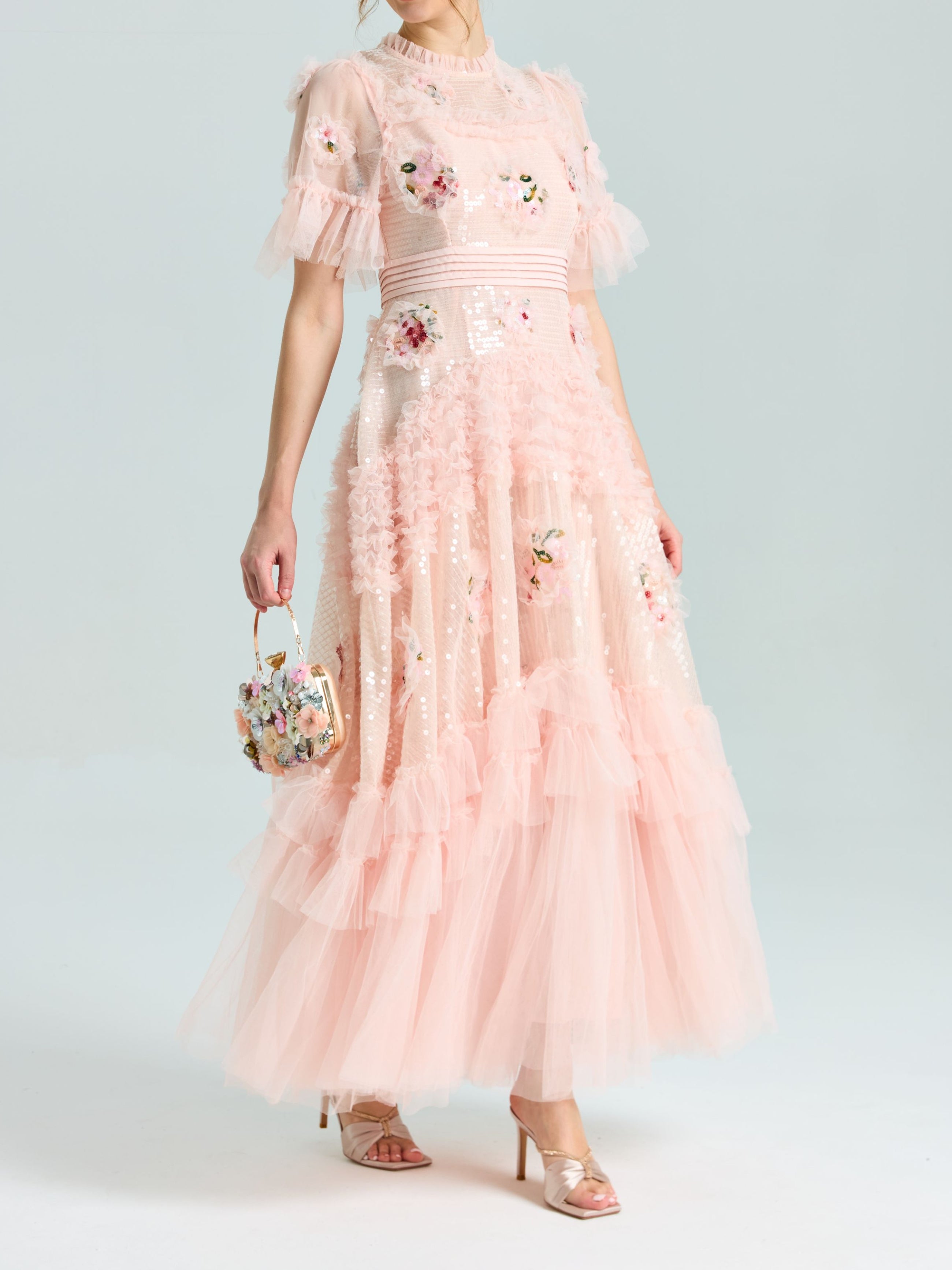 A person wears the Social Queen Embroidered Ruffle Tiered Tulle Dress—light pink with sequin floral embroidery, ruffled layers, paired with a matching floral handbag and open-toe heels—ideal for special occasions.