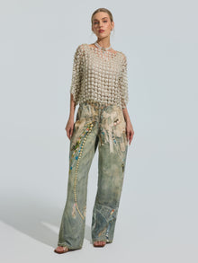 A woman wears the Social Queen Pearl Daisy Cutwork Top (camisole not included), featuring intricate pearl embellishments, paired with high-waisted, wide-leg jeans adorned with colorful beaded designs.