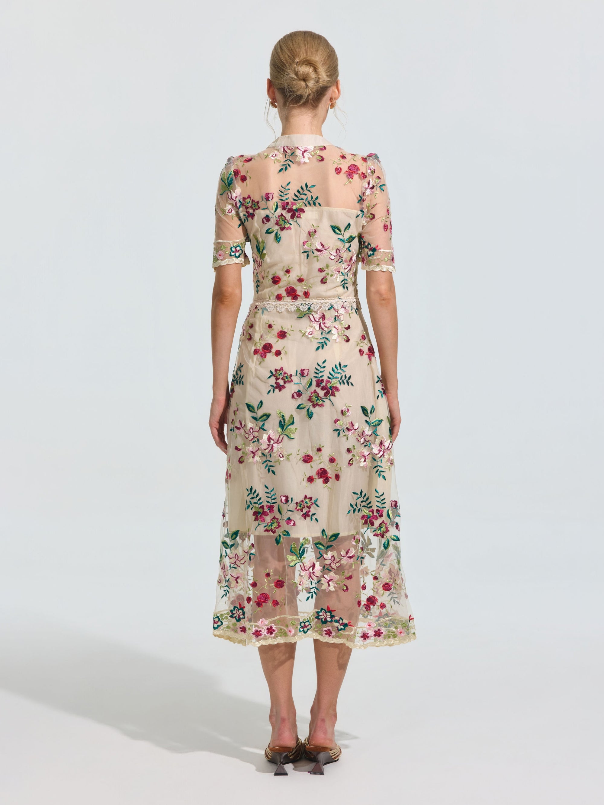 A woman faces away, wearing the Social Queen Floral Embroidered Bow-Tie Dress featuring sheer mesh, an A-line silhouette, and short sleeves, paired with dark heeled sandals.