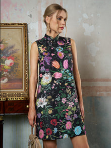 A woman in the Social Queen Colorful Floral Mandarin Collar Dress stands indoors beside a floral painting and a wooden table, holding a beige handbag.