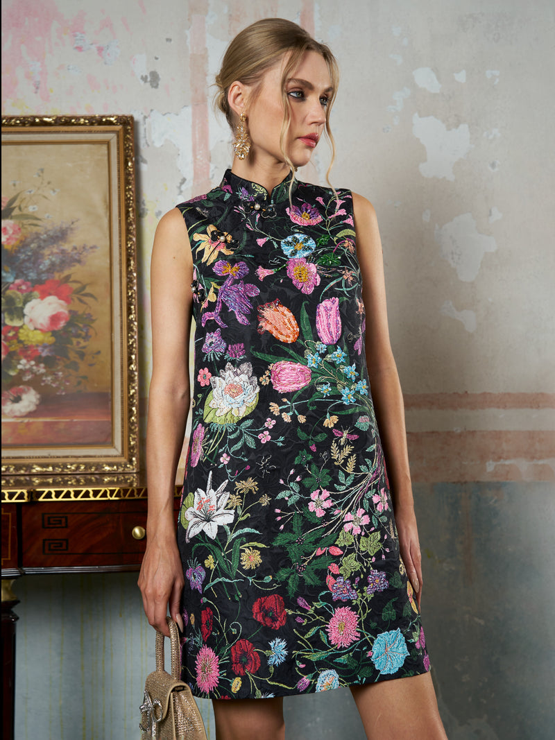 A woman in the Social Queen Colorful Floral Mandarin Collar Dress stands indoors beside a floral painting and a wooden table, holding a beige handbag.