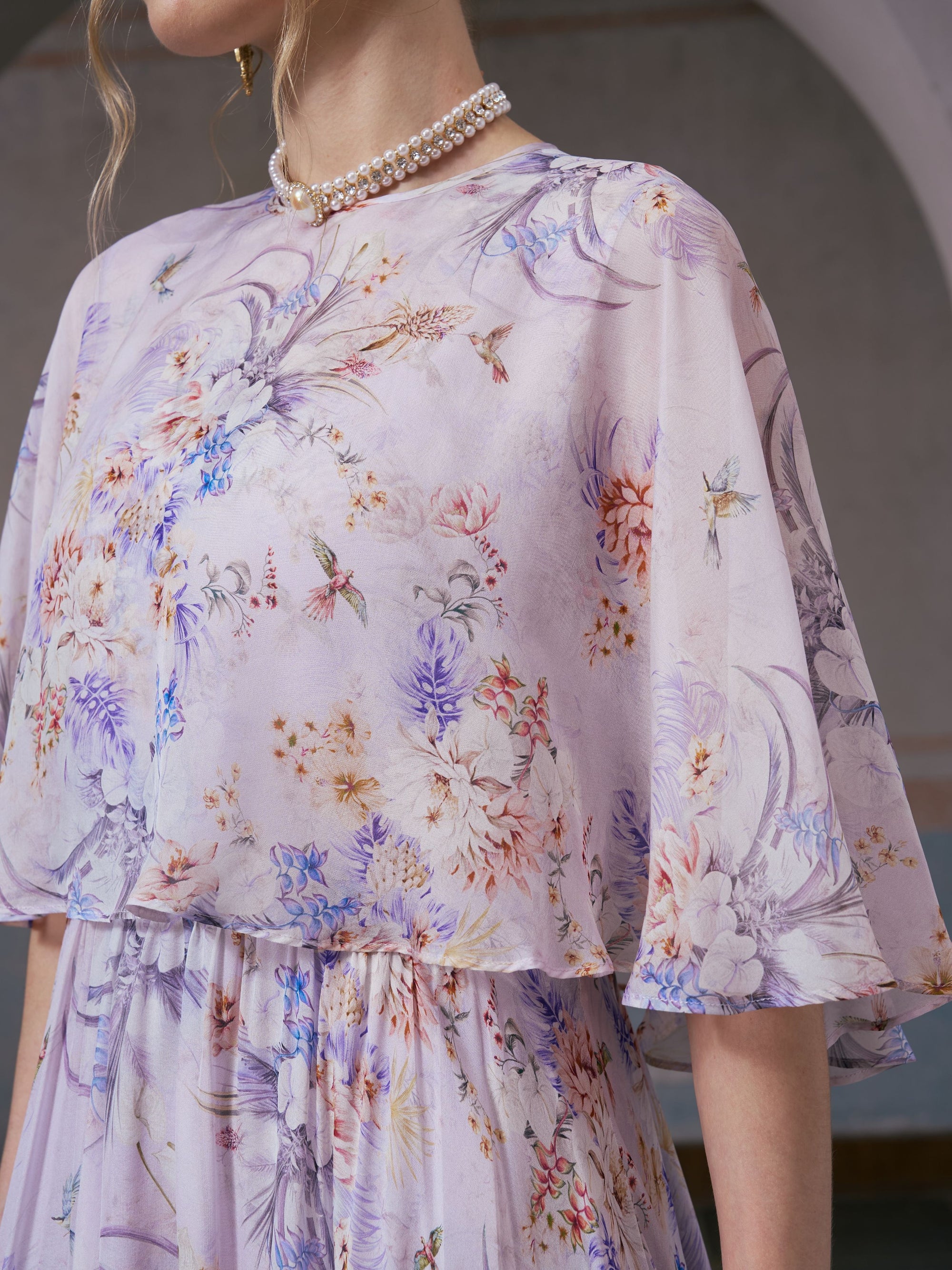 A woman wearing the Social Queen Lavender Print Silk Dress, featuring wide sleeves and a delicate lavender bird motif, paired with a pearl necklace, stands indoors.