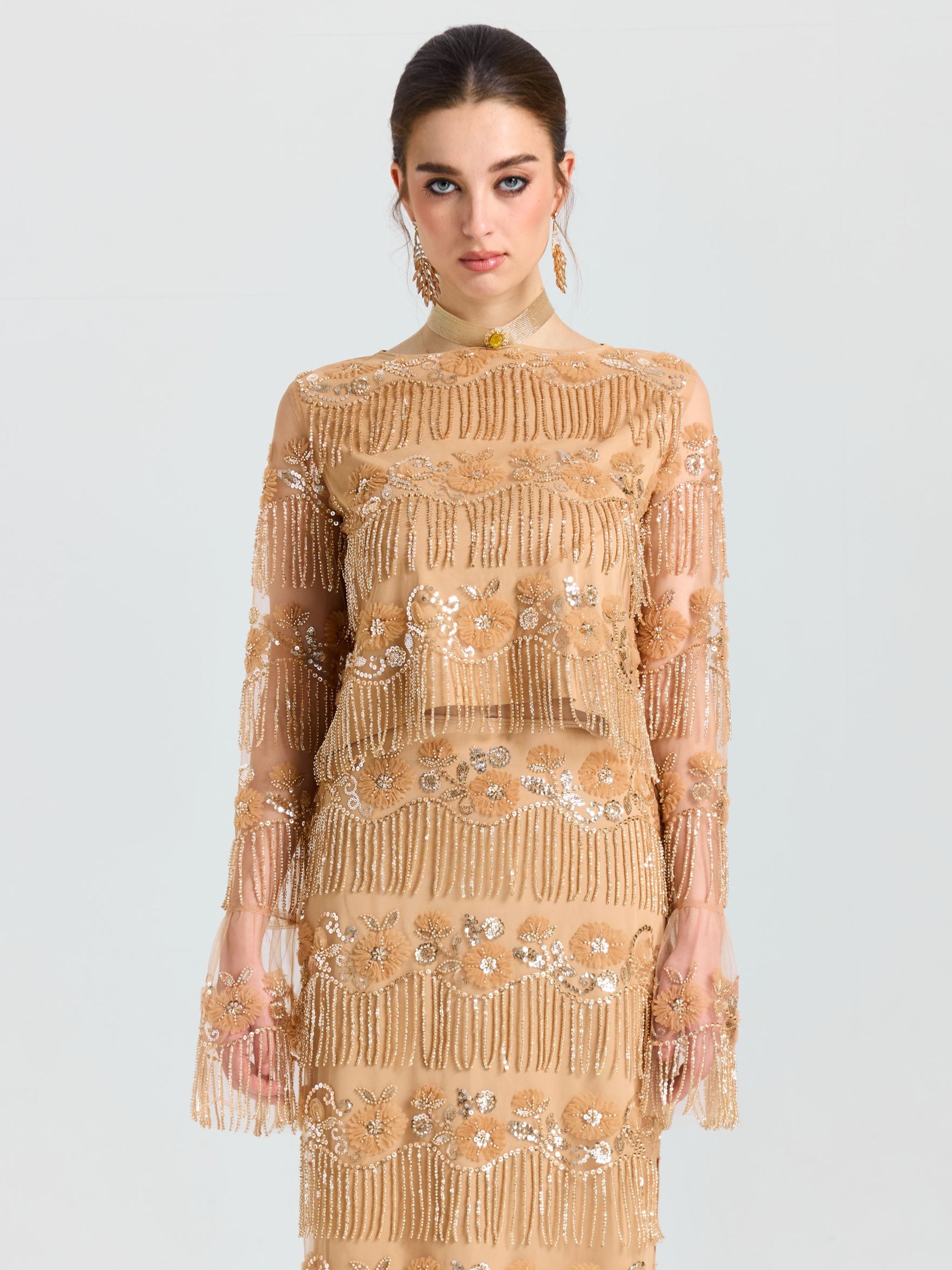 A woman faces forward, wearing the Social Queen Beaded Fringe Floral Top, a long-sleeve sheer mesh piece with floral embroidery and sequin embellishments, set against a plain light background.