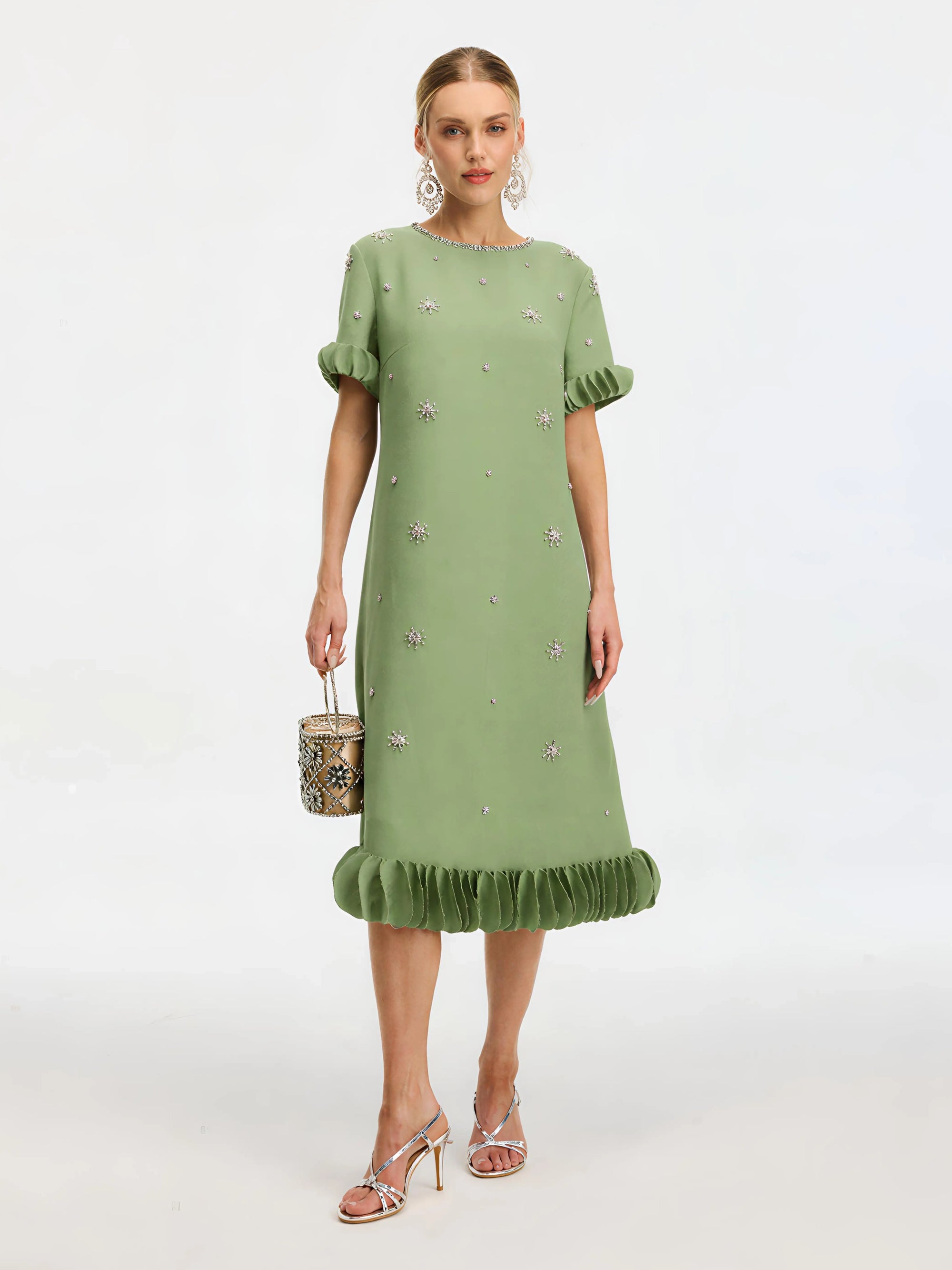 A woman wears the Social Queen Beaded Crystal Embellished Dress, a light green, knee-length shift with ruffled sleeves and hem, crystal accents, and floral details. She carries a handbag and pairs the look with strappy heels.