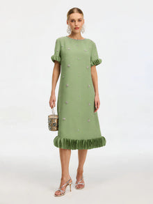 A woman wears the Social Queen Beaded Crystal Embellished Dress, a light green, knee-length shift with ruffled sleeves and hem, crystal accents, and floral details. She carries a handbag and pairs the look with strappy heels.