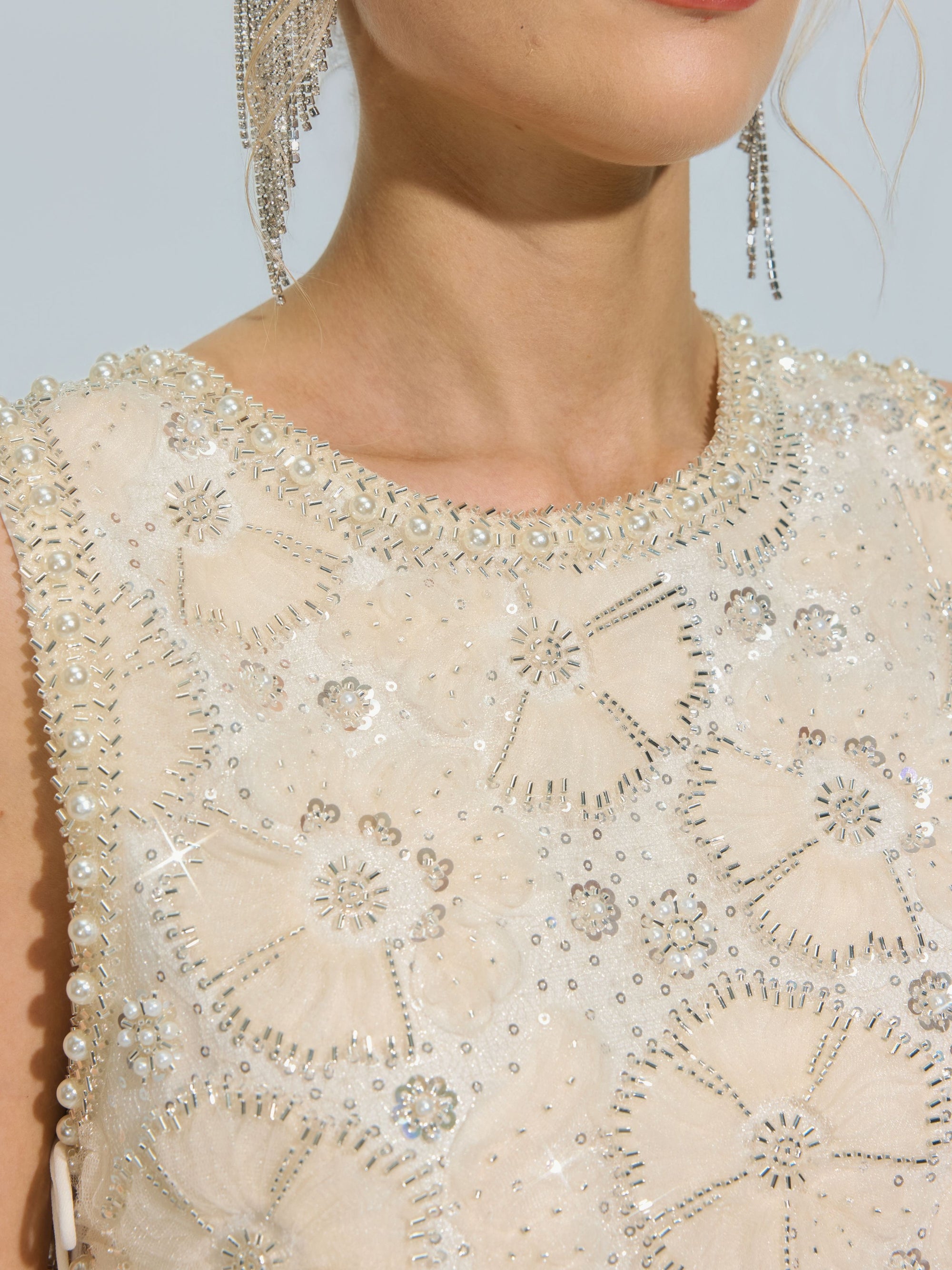 Close-up of a woman wearing the Social Queen Floral Beaded Mesh Sleeveless Top, featuring cream-colored mesh embellished with beaded details, sequins, and rhinestones, paired with long, dangling earrings.