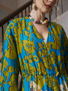 A person wearing the Social Queen Contrast Floral Print Silk Cotton Dress, featuring a v-neckline and blue and yellow floral design, pairs it with a colorful beaded necklace while standing in front of a staircase, epitomizing effortless resort wear style.