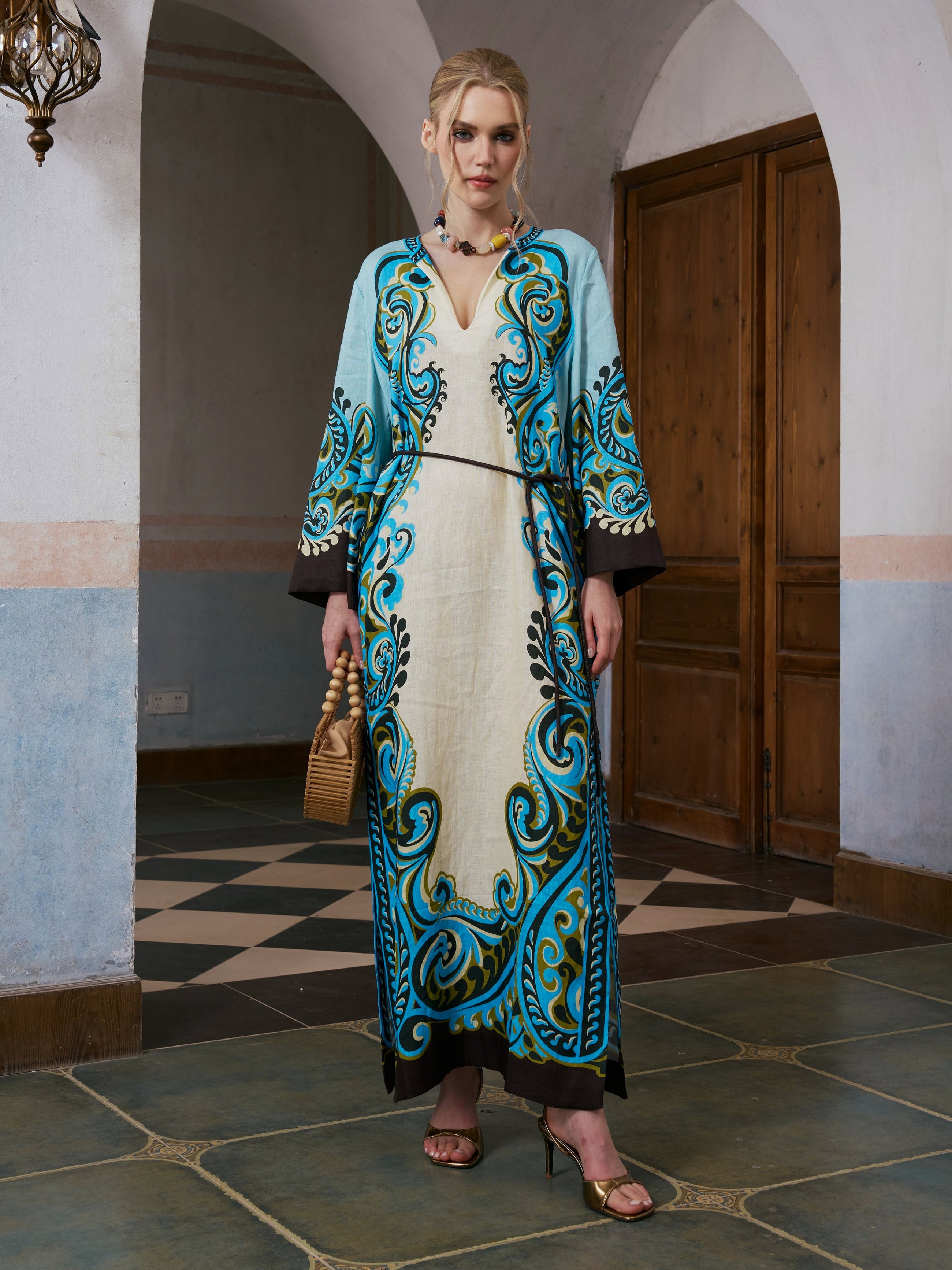 A woman stands indoors wearing the Social Queen Paisley Print Linen Dress with a long, ornate blue and white paisley border print and included belt, accessorized with a small tan handbag and heeled sandals. Wooden doors and a tiled floor are visible in the background.