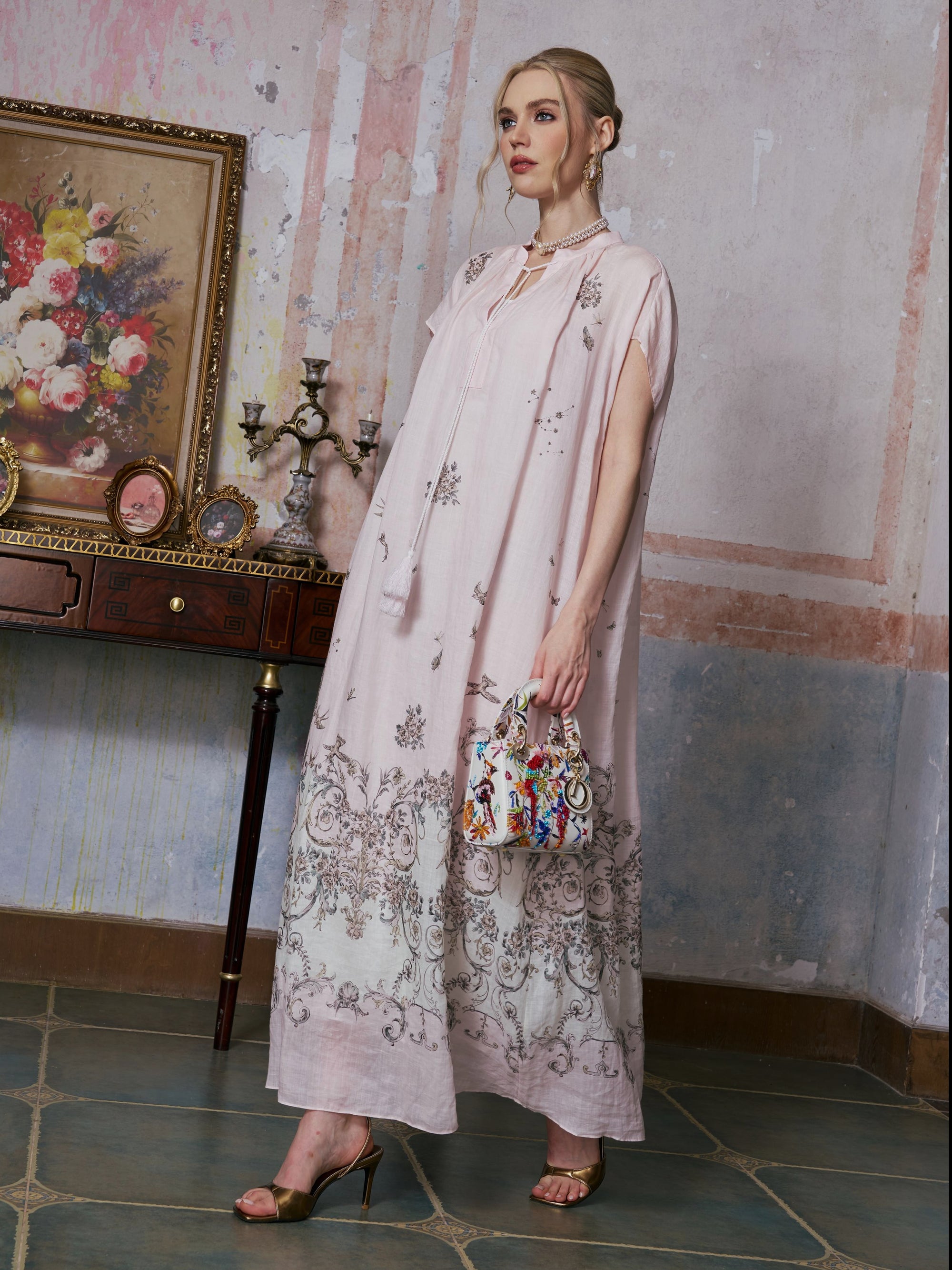A woman wearing the Social Queen Baroque Garden Print Ramie Dress (includes slip dress) holds a colorful handbag as she stands indoors near a table decorated with ornaments and a framed floral painting.
