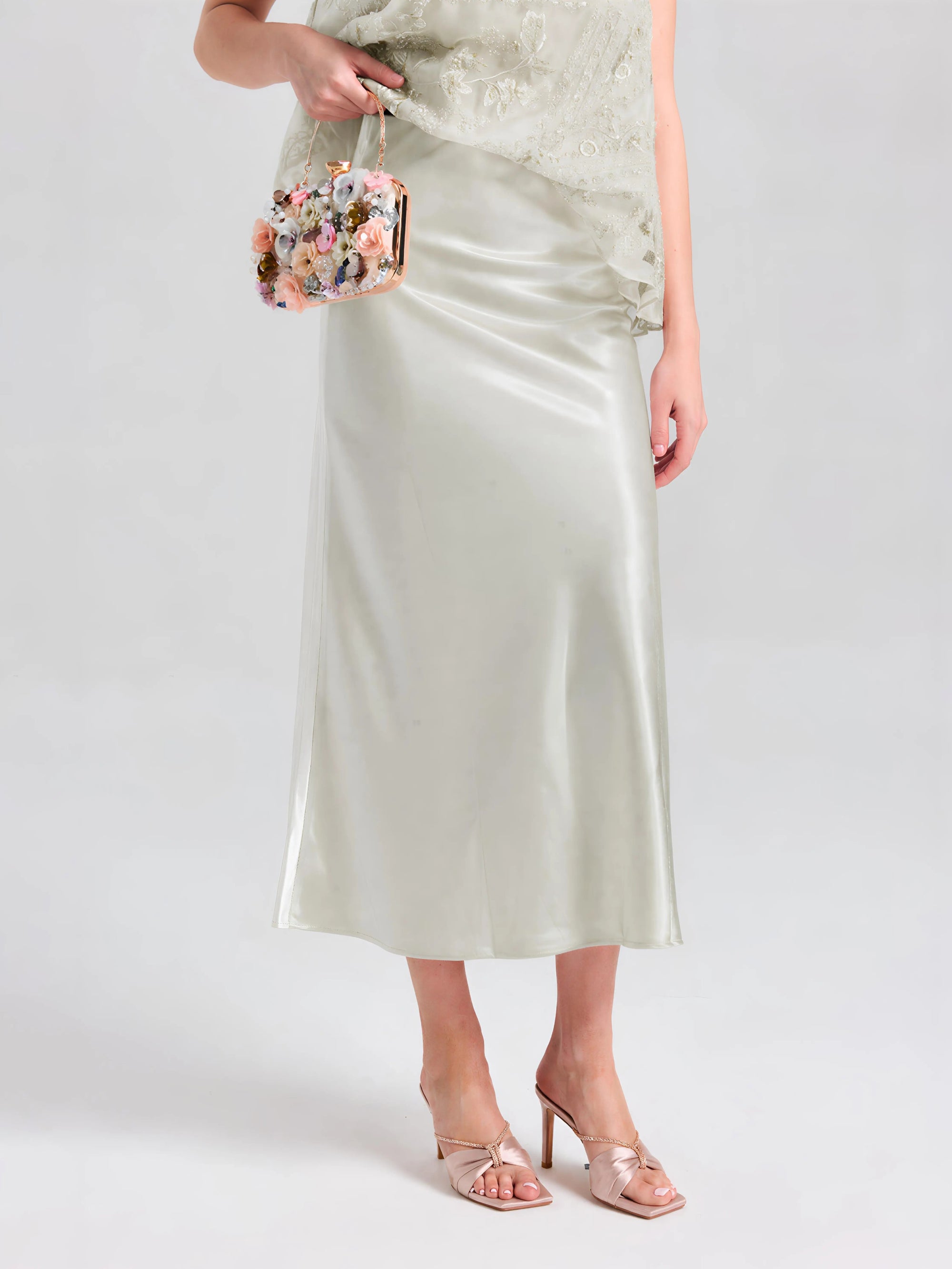 A person wearing the Social Queen Satin A-Line Midi Skirt in pale green with a matching top, open-toe heeled sandals, and holding a small floral-embellished handbag.