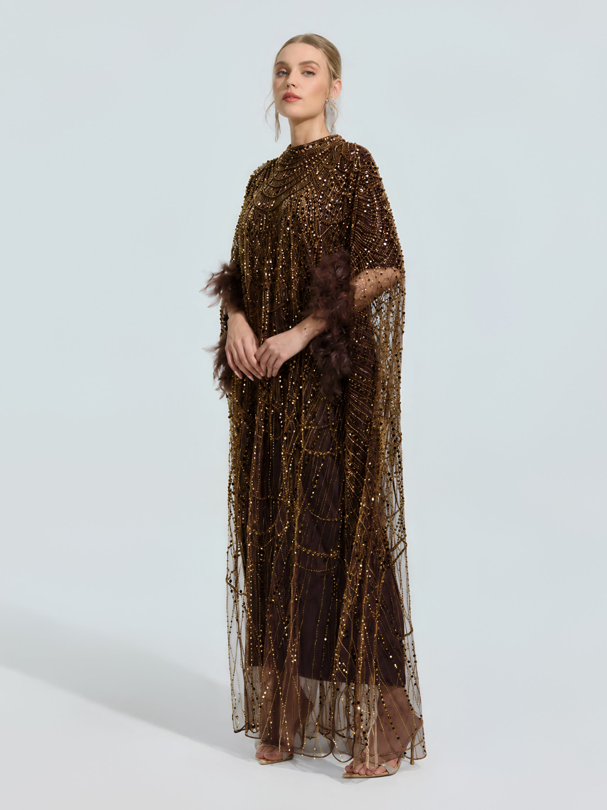 A woman models the Social Queen Beaded Embroidered Feather Trim Cape Gown, a floor-length brown evening dress with feathered sleeves, against a plain light background, styled with open-toe heels.