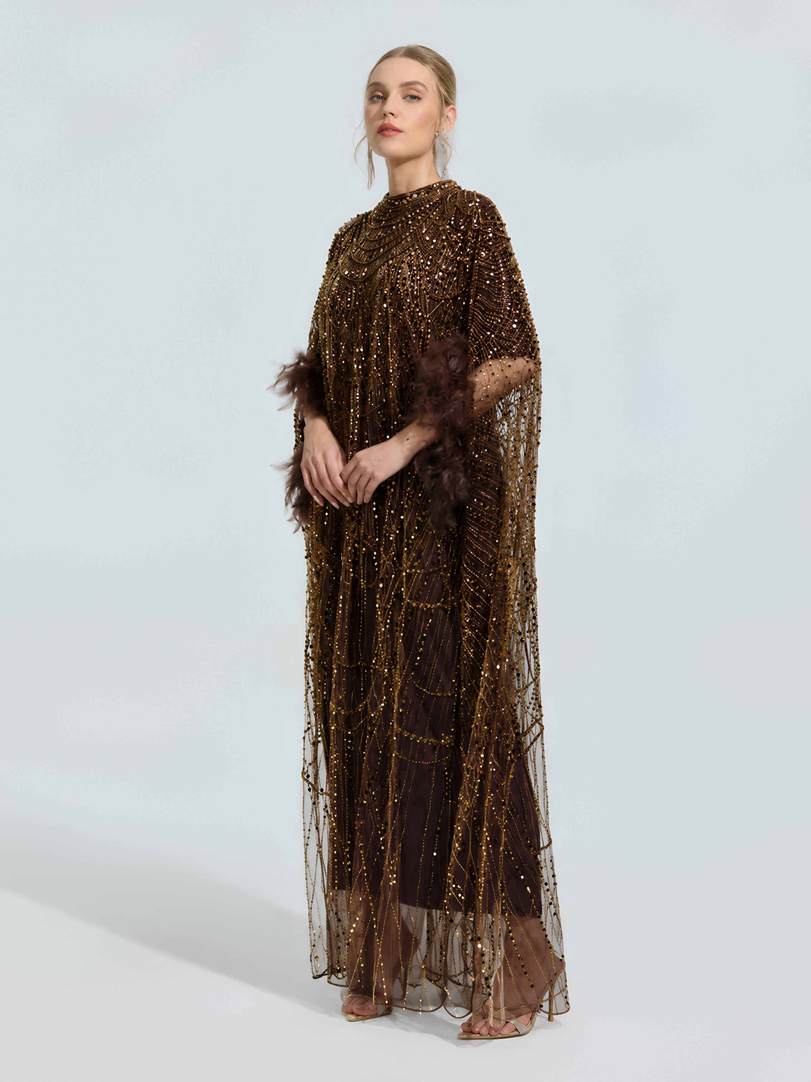 A woman models the Social Queen Beaded Embroidered Feather Trim Cape Gown, a floor-length brown evening dress with feathered sleeves, against a plain light background, styled with open-toe heels.