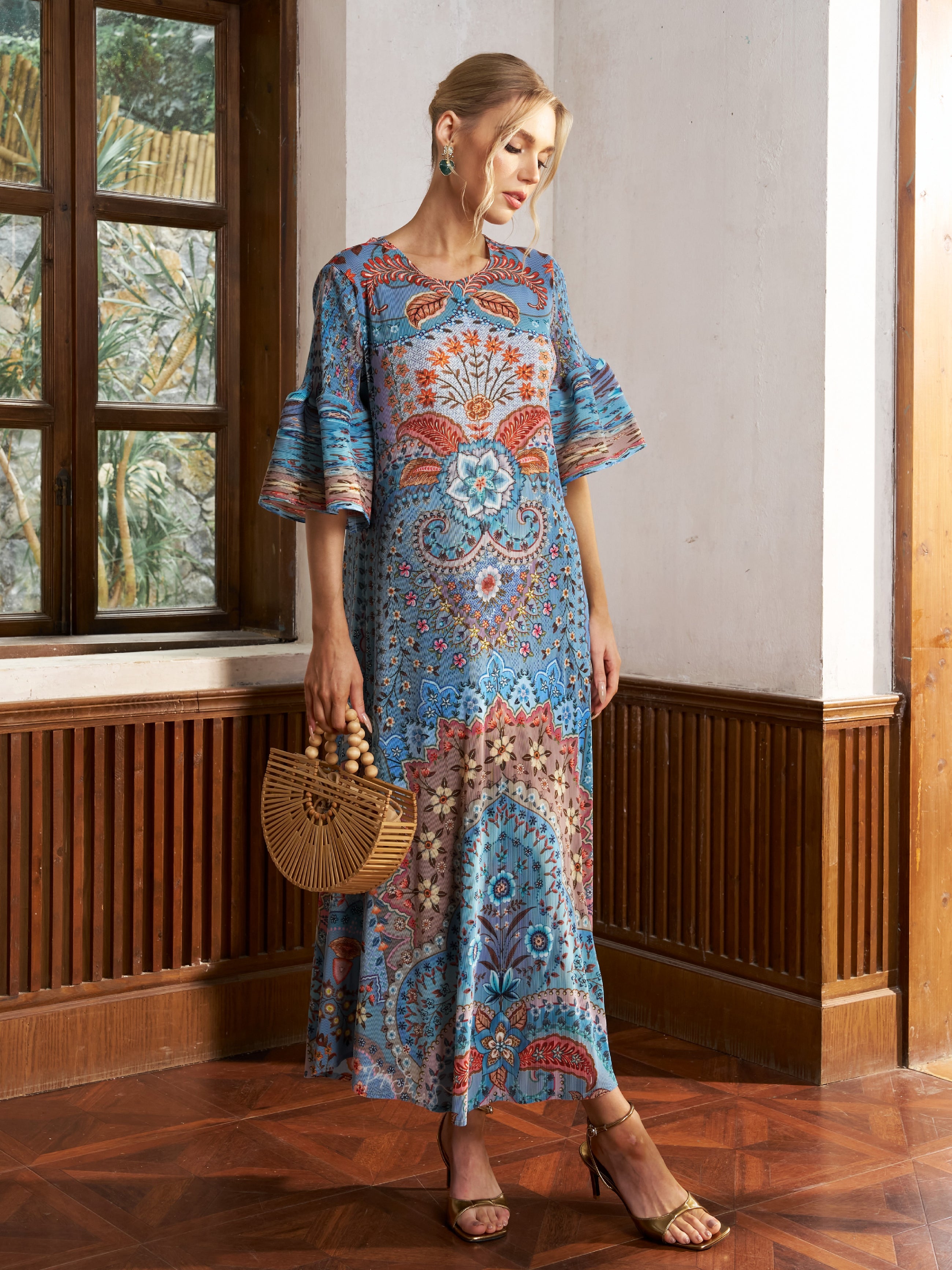 A woman in the Social Queen Paisley Print Pleated Midi Dress stands indoors, pairing the flared-sleeve blue dress with a wooden handbag, sandals, and earrings for an effortless vacation look.