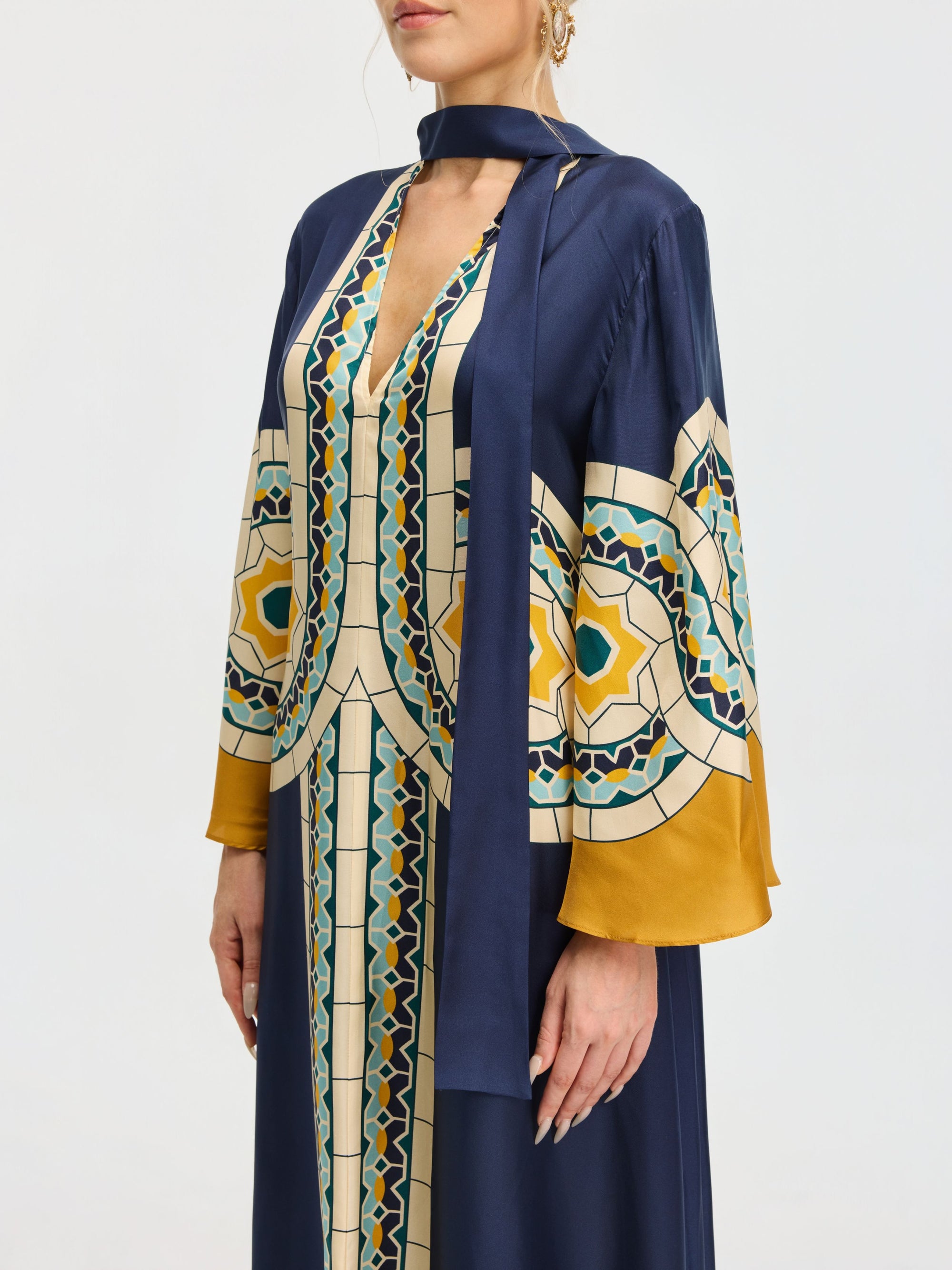 A person wearing the Social Queen Moroccan Print Silk Caftan Dress, featuring a long navy blue silhouette with wide sleeves, geometric yellow, teal, and cream patterns, and a matching neck scarf.
