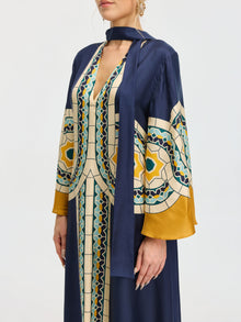 A person wearing the Social Queen Moroccan Print Silk Caftan Dress, featuring a long navy blue silhouette with wide sleeves, geometric yellow, teal, and cream patterns, and a matching neck scarf.