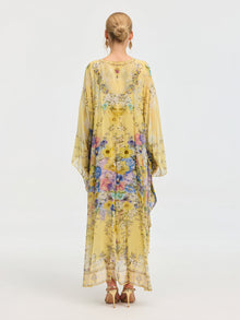 A woman in the Social Queen Floral Print Silk Caftan Dress, featuring wide sleeves and a yellow floral pattern, stands with her back to the camera. Her hair is in a neat bun, accessorized with high heels and hoop earrings.
