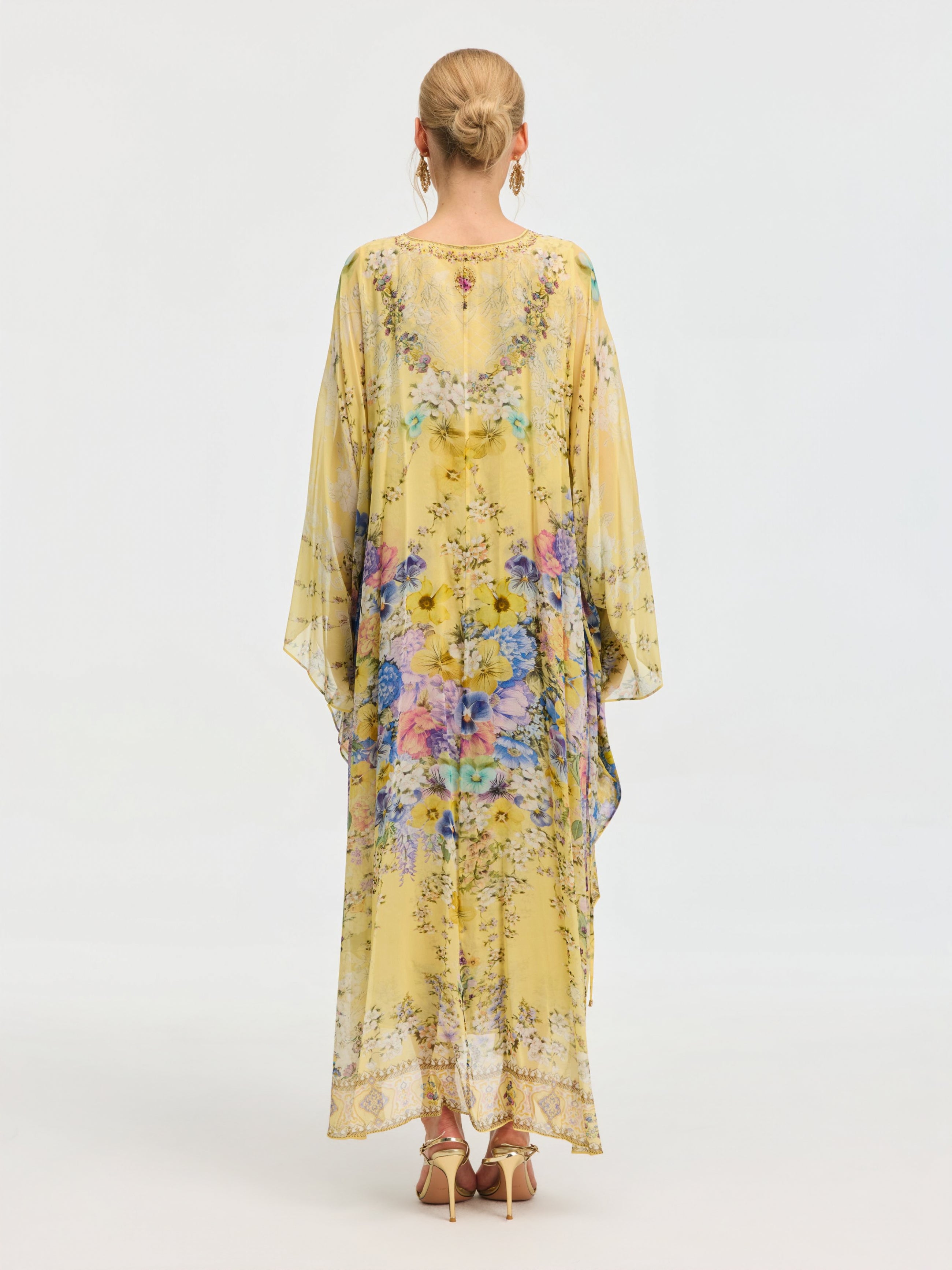 A woman in the Social Queen Floral Print Silk Caftan Dress, featuring wide sleeves and a yellow floral pattern, stands with her back to the camera. Her hair is in a neat bun, accessorized with high heels and hoop earrings.