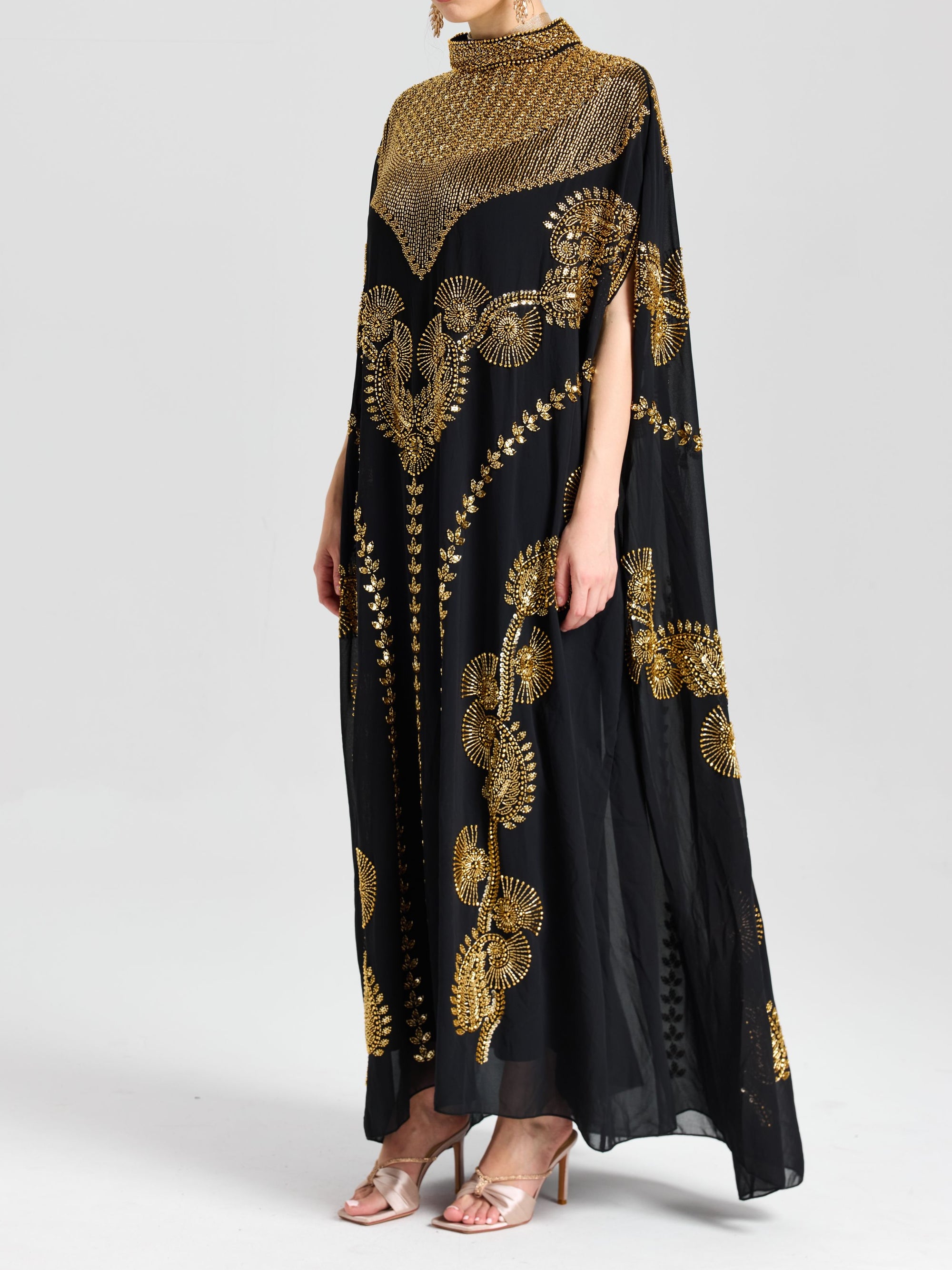 A person wears the Social Queen Beaded Embroidered Cape Dress—long, black, with intricate Persian-inspired gold embroidery—standing against a plain light background.