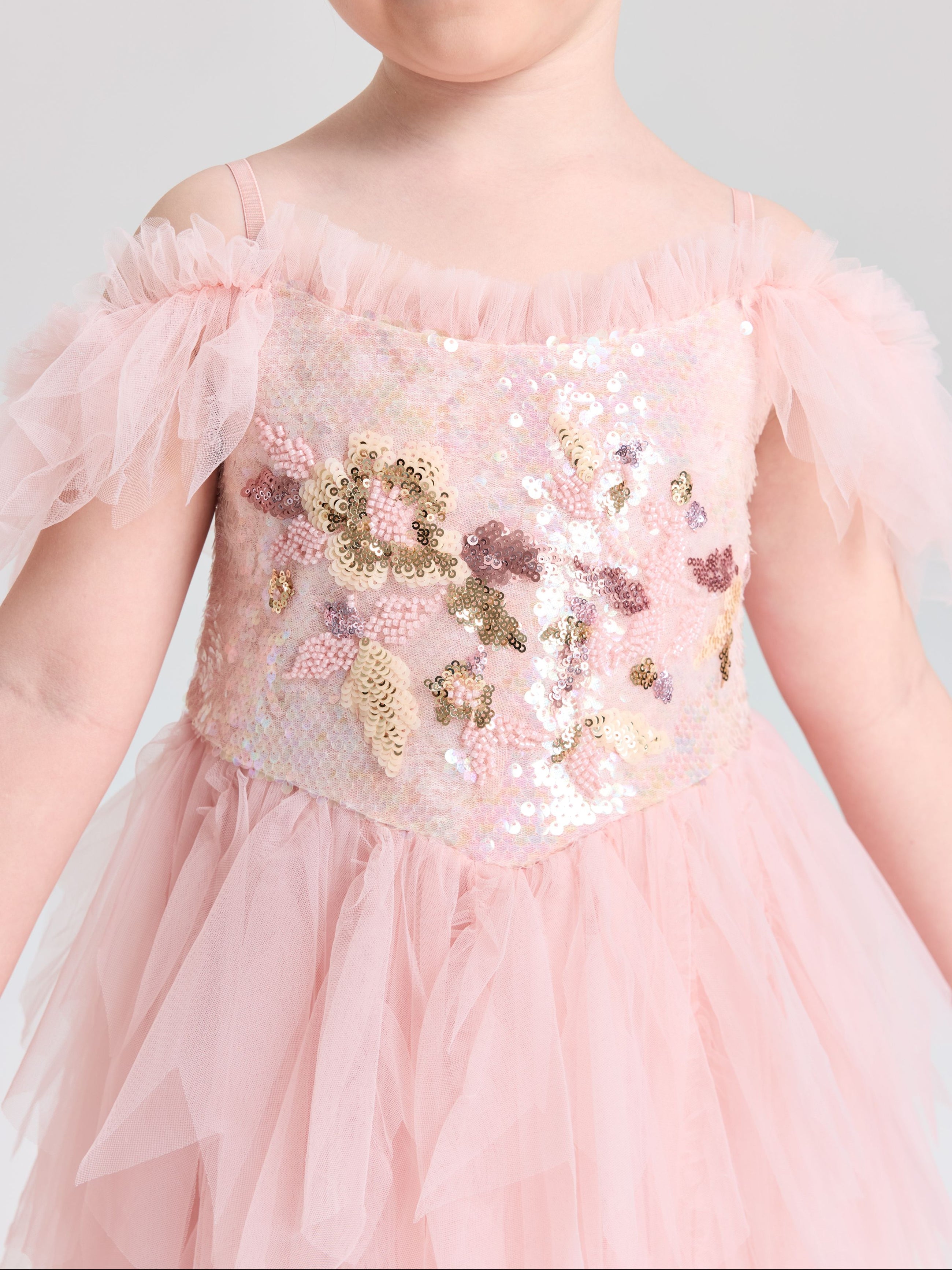A young child wears the Social Queen Girls Floral Sequin Tulle Dress, featuring light pink tulle sleeves and gold, pink, and silver embellishments on the floral sequin bodice.