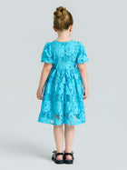 A young girl faces away in the Social Queen Girls 3D Floral Appliqué Cutwork Lace Dress featuring short sleeves and black shoes, set against a light gray background.